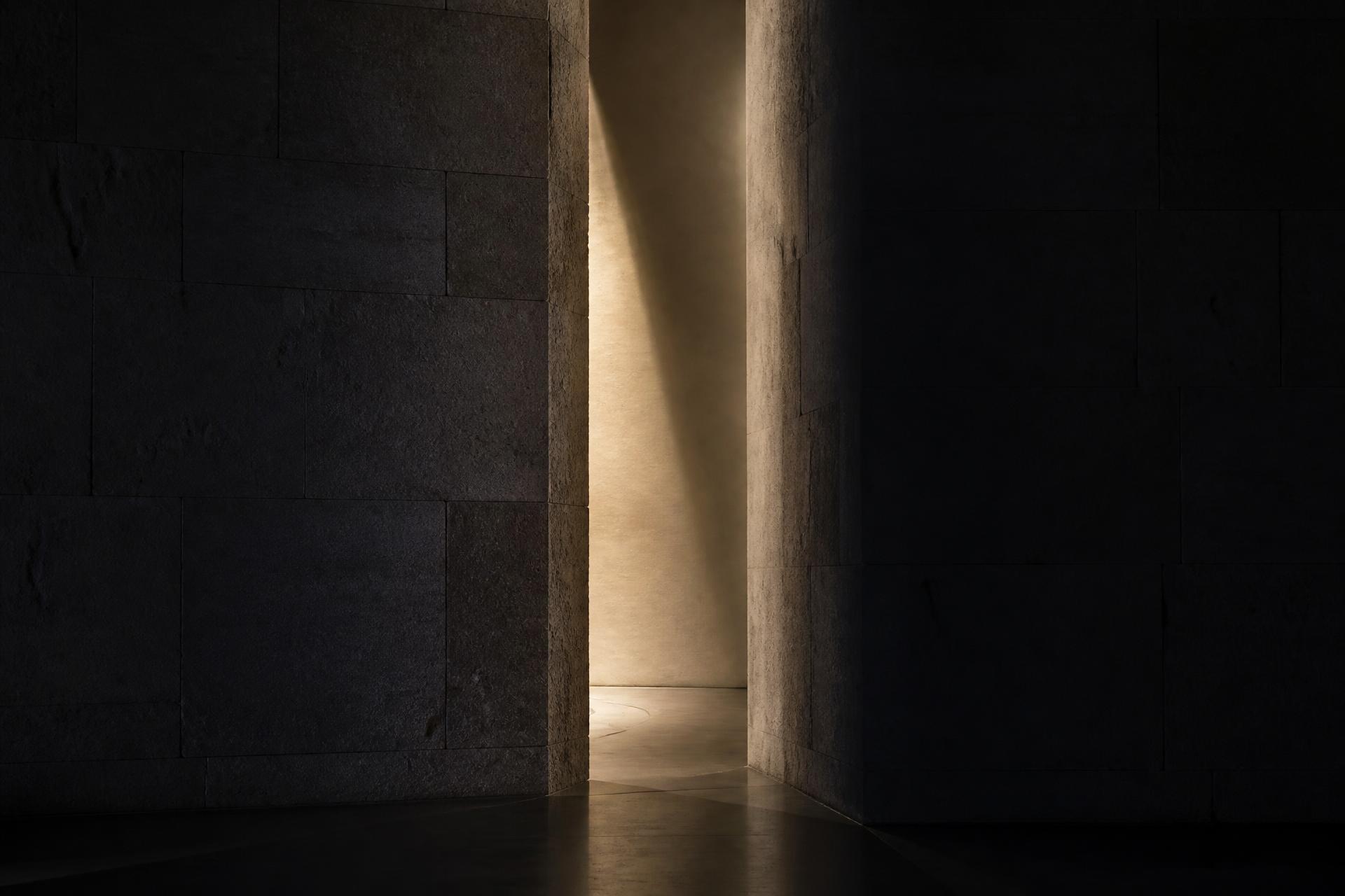 Fenda de luz atravessando uma parede de pedra escura — o limiar entre dois estados.